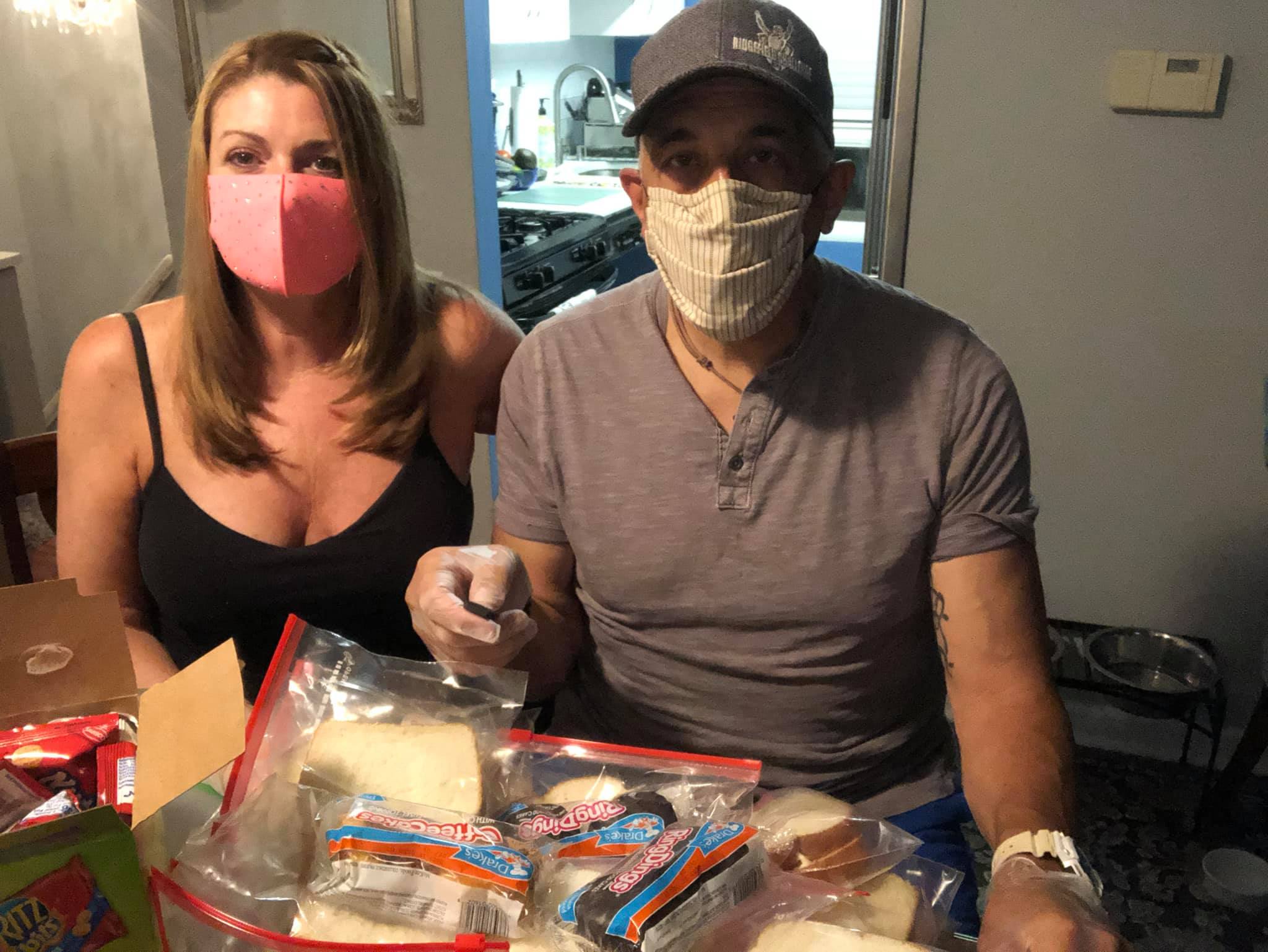 A couple posing in masks in front of some bagged sandwiches and snacks